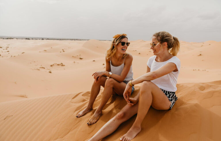 Cheerful Friends Sitting On Sand At Desert Against Sky stock photo